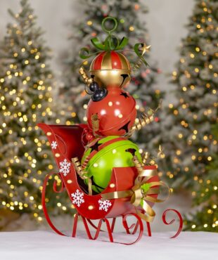 The item is a small decorative red sleigh filled with three large, shiny jingle bells stacked on top of each other like a snowman. The bottom bell is green, the middle one is red with stars, and the top one is gold and red. The sleigh has white snowflakes painted on it and a gold ribbon on the front.